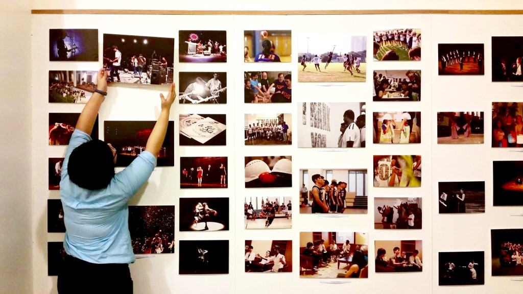 A person wearing a blue shirt is putting up a photograph print of a band performing on a wall full of photograph prints.
