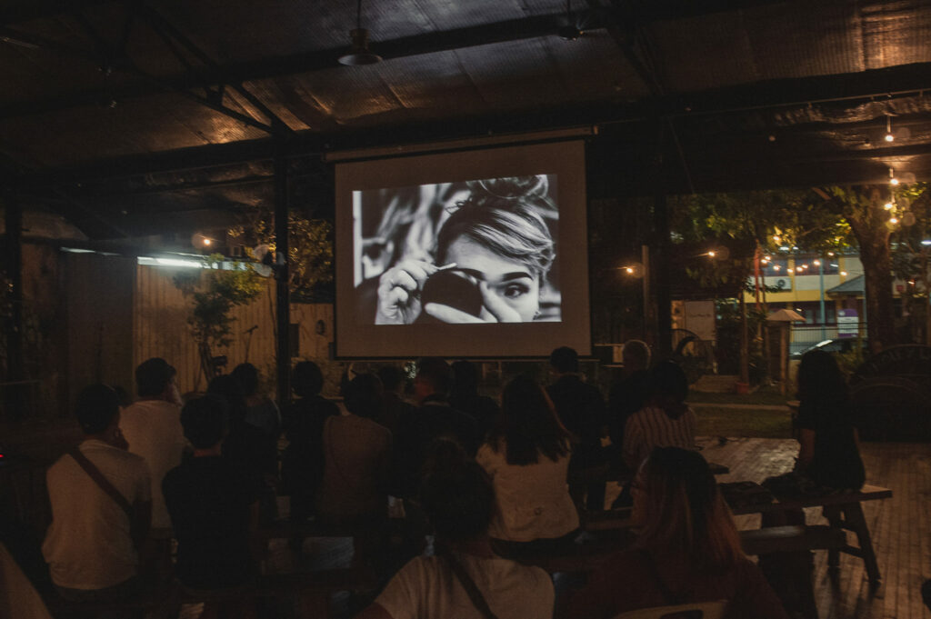 A projector showing a film on screen where people are seated and watching in an open space at night time.