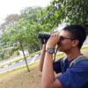 A portrait of Golam Rabbani who has short black hair, wearing glasses and blue t-shirt, using a binoculars with street and trees in the background.