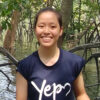 A headshot of smiling Ning Yiran who has a long black hair in pony tail, wearing a blue t-shirt that says “Youth Expedition Project” with mangrove forest and water surface in the background.