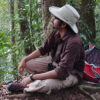 A portrait of Vinod Kumar Saranathan who is sitting sideways on the ground, looking slightly up and staring into space, wearing a white hat, a sunglasses, brown t-shirt, and beige pants with a forest in the background.