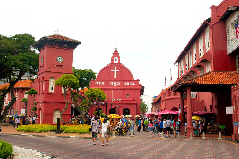 A landscape of red-painted church named Christ Church Melaka, an Anglican church in Malacca City, Malaysia with a red-painter clock tower next to it, and many visitors walking around the area.  