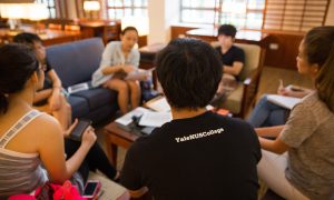 A group of students sitting in a circle in a group discussion at the library’s level 1 study area couches with one student is being pictured from the back, wearing the Yale-NUS College black shirt.