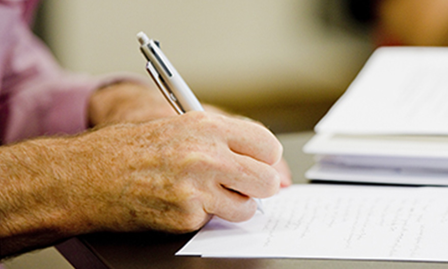 A close-up of someone’s hand holding a pen, writing on a piece of paper on a table.
