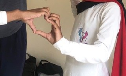 A person who wears a white long-sleeve shirt forms one side of heart hand-gesture and another person who wears a grey top forms the other side of heart hand-gesture, forming a complete hand gesture in a room.