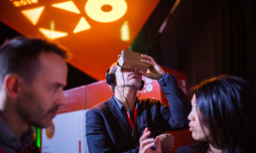 A man who wears a blue suit using the Virtual Reality (VR) headset and holding them, and there are two people having conversations in front of him.