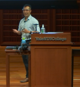 A person standing behind the “Yale-NUS College” podium, talking to the audience and presenting at the Yale-NUS Performance Hall. 