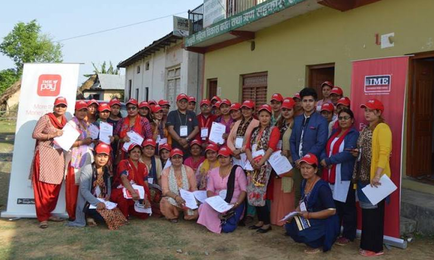 A group of people wearing IME red cap, holding a paper with the IME pay standing banner on the left of the group and the IME standing banner on the right of the group with houses in the background.