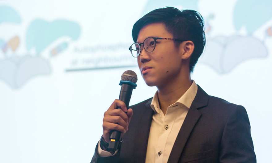 A close-up of a student who has short black hair, wearing a black suit and glasses, holding a microphone, talking to the audience with a presentation in the background.