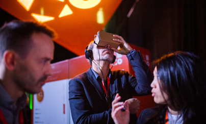 A man who wears a blue suit using the Virtual Reality (VR) headset and holding them, and there are two people having conversations in front of him. 