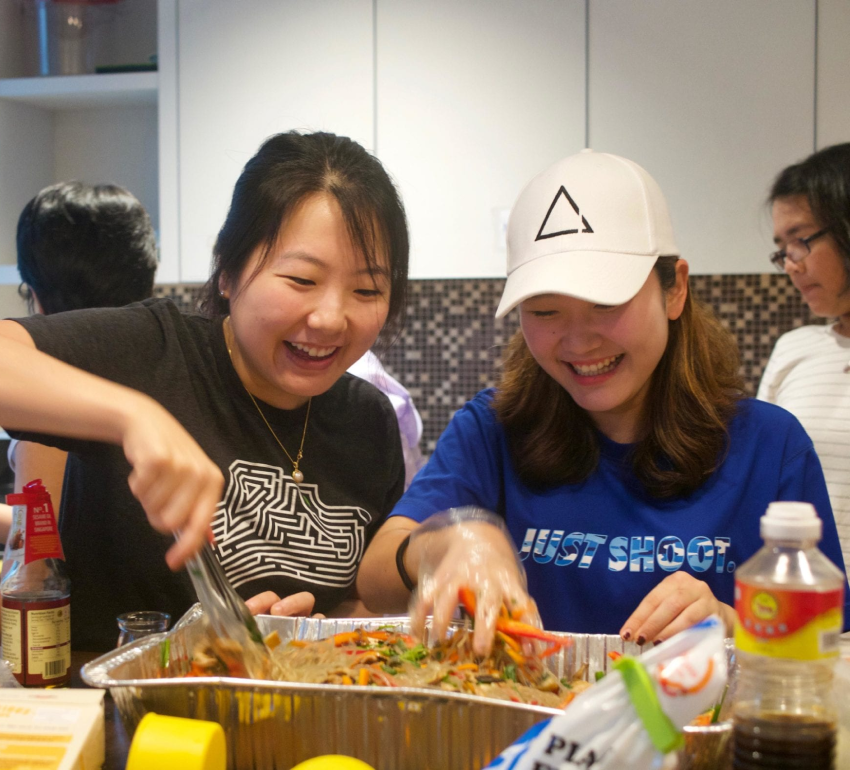 Two students happily mixing food ingredients on a disposable aluminium foil tray with other food ingredients next to it and other people in the background at the Cendana Buttery. 