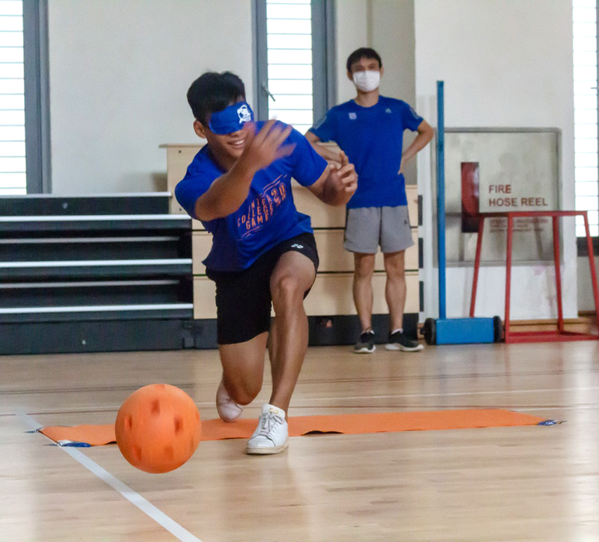 A student playing goalball, wearing an opaque eyeshades, at a position in throwing the orange ball at the Yale-NUS Multipurpose Hall and a person watching in the background.