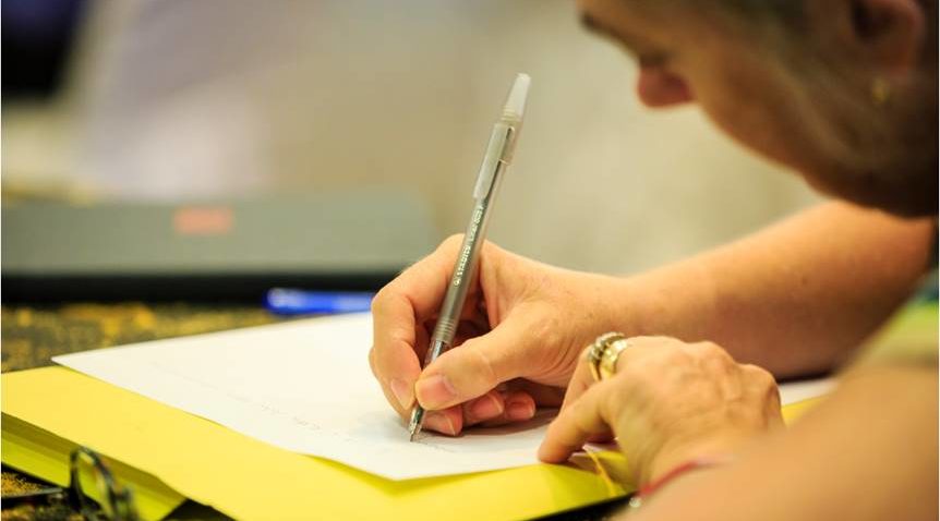 A close-up of a hand of a person writing on a piece of paper with a black pen on top of a yellow surface