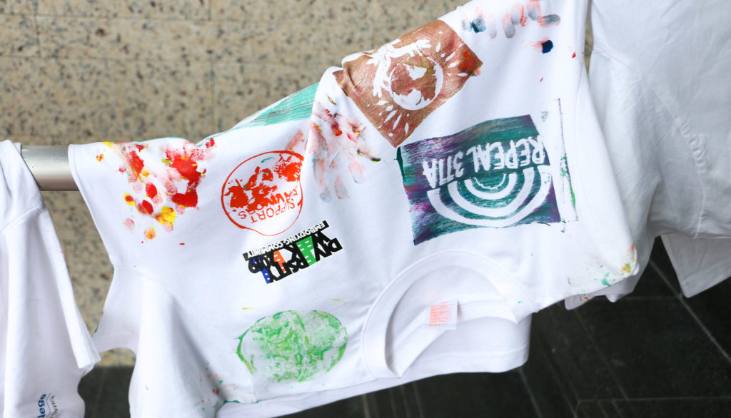 A white t-shirt with some diversity week 2022 paintings and artworks, hanging on a handrail. 