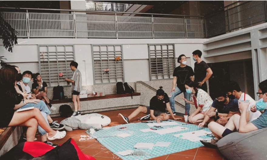 A group of students who wear masks are gathering, drawing, and coloring at the open space below Cafe Agora stairs, between Yale-NUS Multipurpose Hall and the Library. 