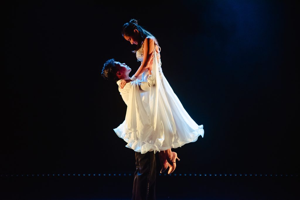 Two students performing ballroom where a male student wearing white t-shirt and black pants are lifting the other female student who wears white dress during a ballroom performance at the Yale-NUS Black Box Theatre.
