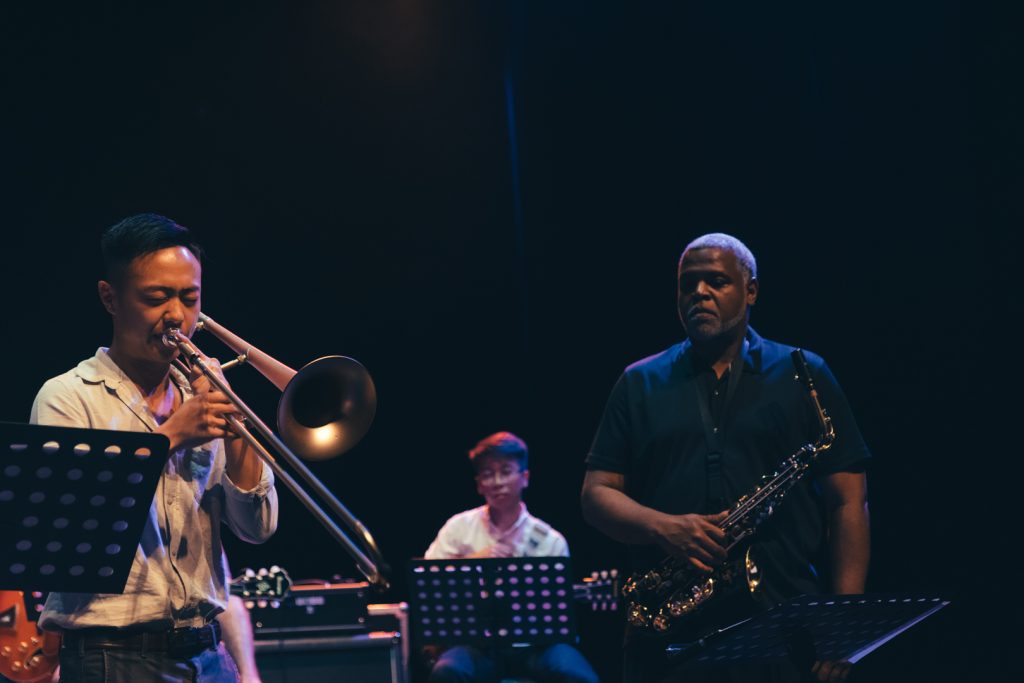 Three musicians performing on stage at Yale-NUS Black Box Theatre, where one is playing a trombone on the left, one is playing a saxophone on the right, and the other one at the back is playing a guitar.