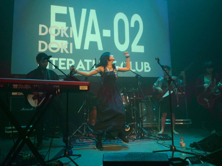 A band performing on stage during the “Bread and Jam” concert where the singer is seen jumping around on stage with three guitarists on stage and a screen showing visualization saying “Doki-Doki” in the back at Yale-NUS Black Box Theatre.