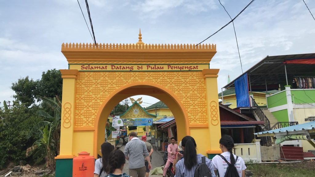 A landscape of yellow-painted concrete gate with traditional patterns at the front of a village that says “Selamat Datang di Pulau Penyengat” which translates to “Welcome to Penyengat Island” in Bintan, Indonesia. 