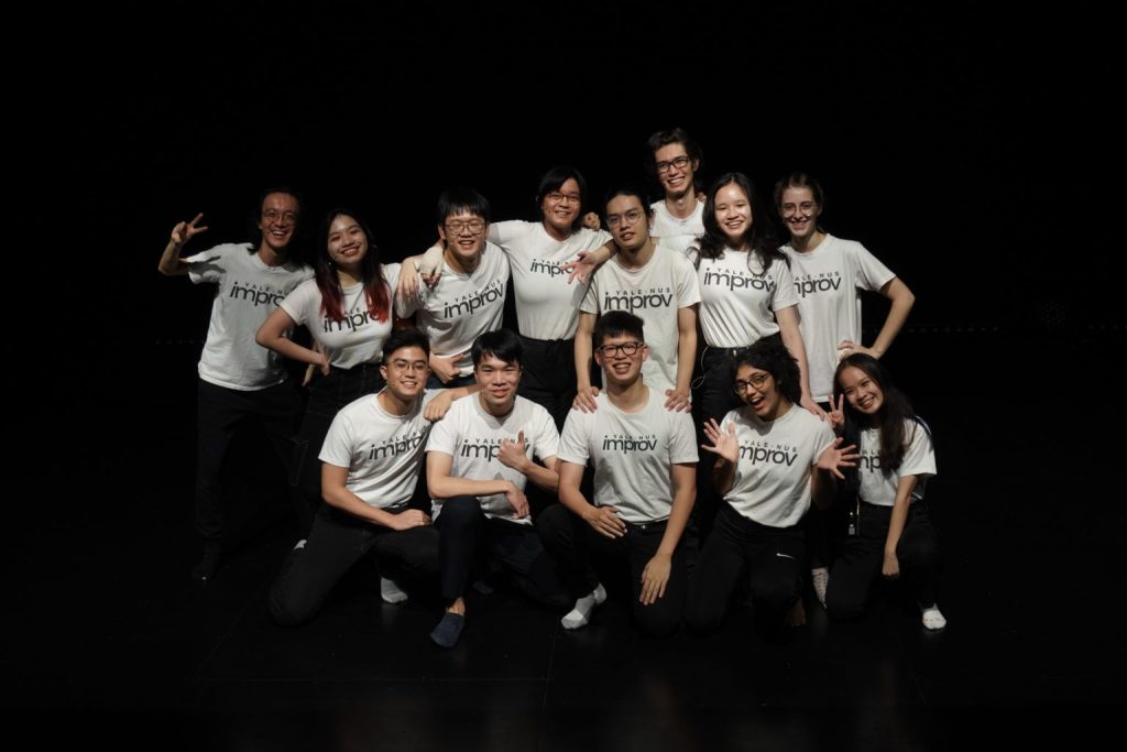 A group of students wearing a Yale-NUS Improv white t-shirt, posing for a picture at the Yale-NUS Black Box Theatre.
