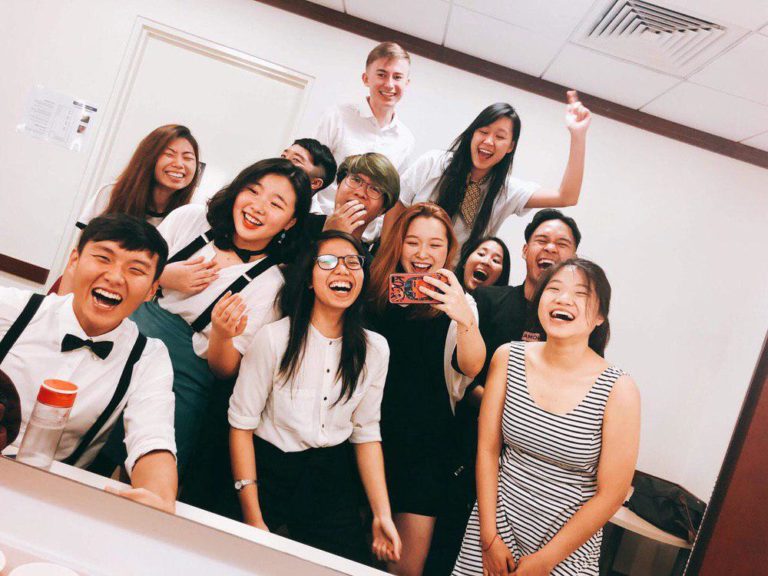 A group of students wearing black-white outfit posing for a picture in front of a mirror, where one of them is taking a “wefie” at the changing room of Yale-NUS Black Box Theatre.