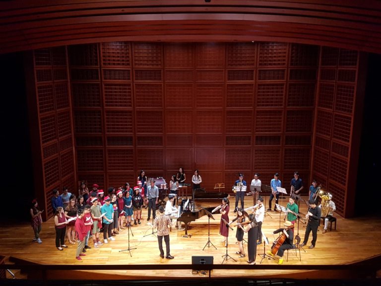 A nosebleed view of the Yale-NUS Chamber Music Collective performing for a christmas concert at the Yale-NUS Performance Hall Theatre, which includes a group of students singing in a choir on the left stage, a pianist on the center stage, a conductor on front stage, and an orchestra on the right stage.
