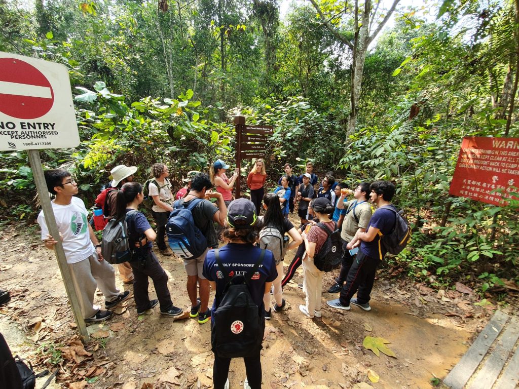 A portrait of group of students on a Learning Across Boundaries (LAB) expedition, wearing backpacks, forming a circle for a briefing, with forest in the background, a “No entry” sign on the left side and “Warning” red sign on the right side.  