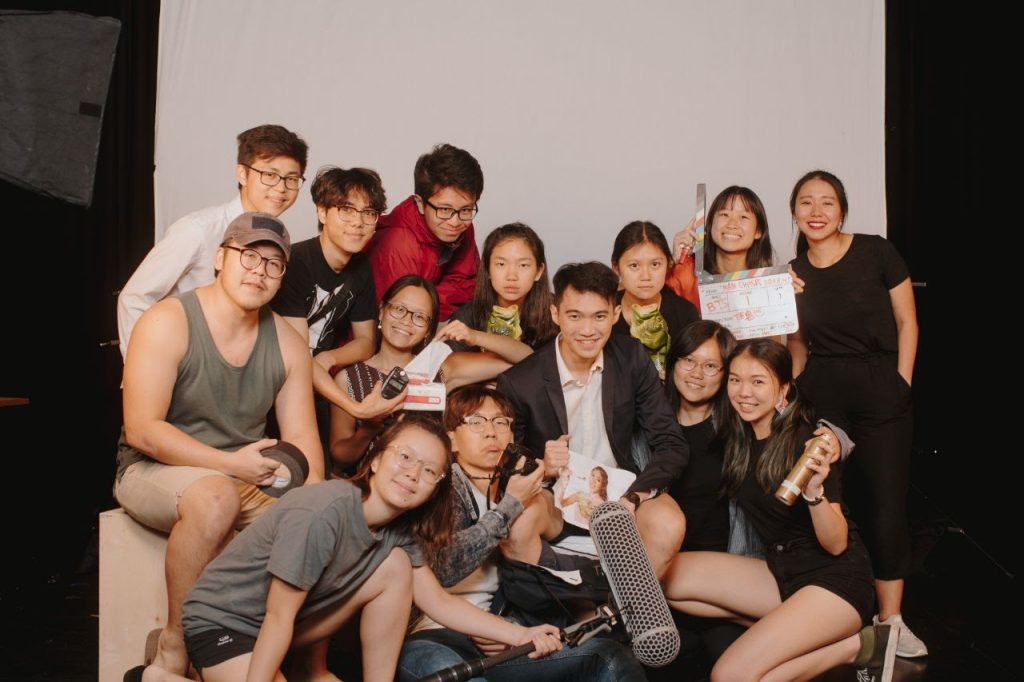 A group of students, each holding a different filming production properties like a clapper, a microphone boom, posing for a picture in front of a white wallpaper backdrop with a studio photography softbox light kit on the left side.  