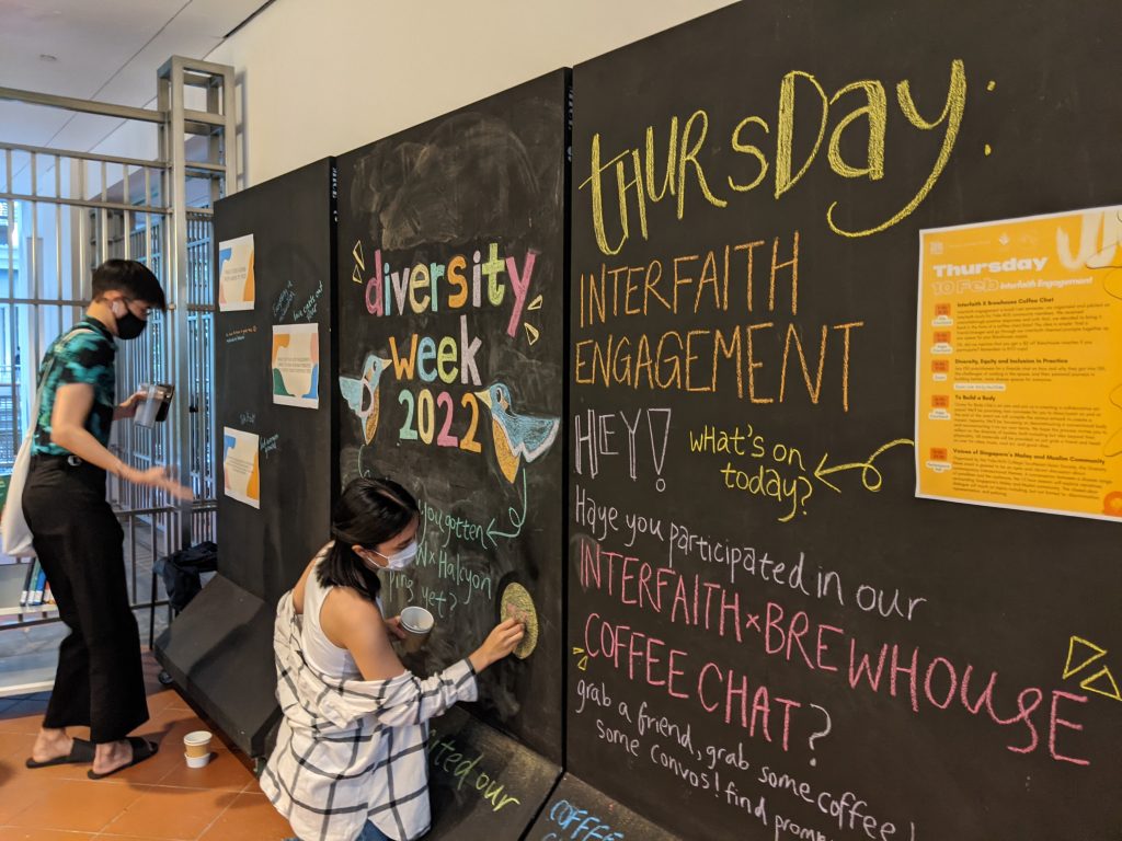 The three black chalk boards at the Elm College gate, showing the Diversity Week 2022 chalk arts and schedule with a student squatting to draw an art with her chalk on one board and the other student looking at the schedule on the other board. 