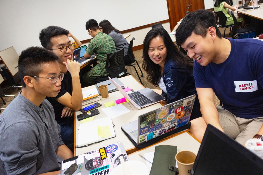 A group of 4 students working together, sitting in opposite sides of the table with papers and laptops on the table, and they’re looking at one laptop at the side of the table in a classroom where there are other students. 