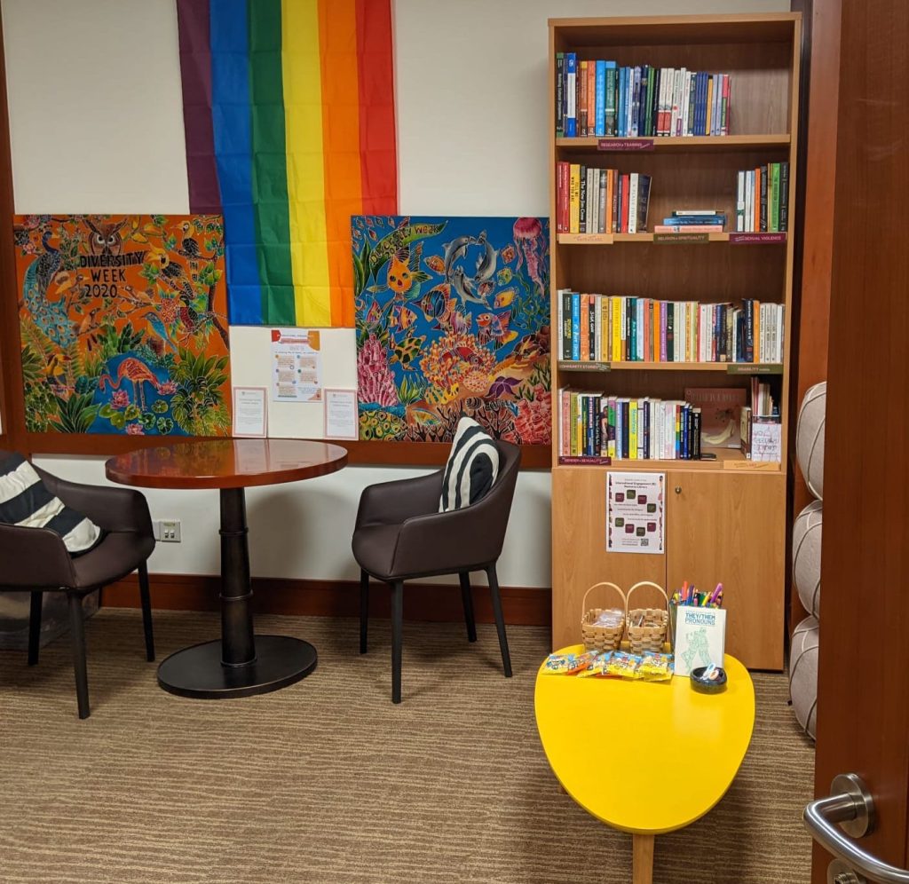 The Intercultural Engagement Space where it has two chairs that has a black-white cushion each with a round table in the middle of it, a pride flag hanging on the wall and two artworks, a shelf full of books next to it, and a small yellow coffee table with stationaries and snacks on top, located at the Student Affairs Office. 