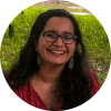 A headshot of smiling Tanya Sharma who has long black hair past her shoulders, wearing a red polkadot top and glasses with black frame with green grass in the background.