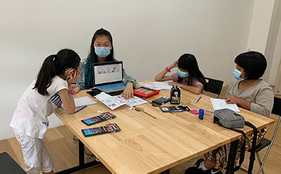 Huanyan, from the Class of 2023, is sitting down on a chair in front of the table full of notebooks, drawings, and stationaries with three students around where she holds a laptop with comic artwork on display, facing the students at the table where the students listen.