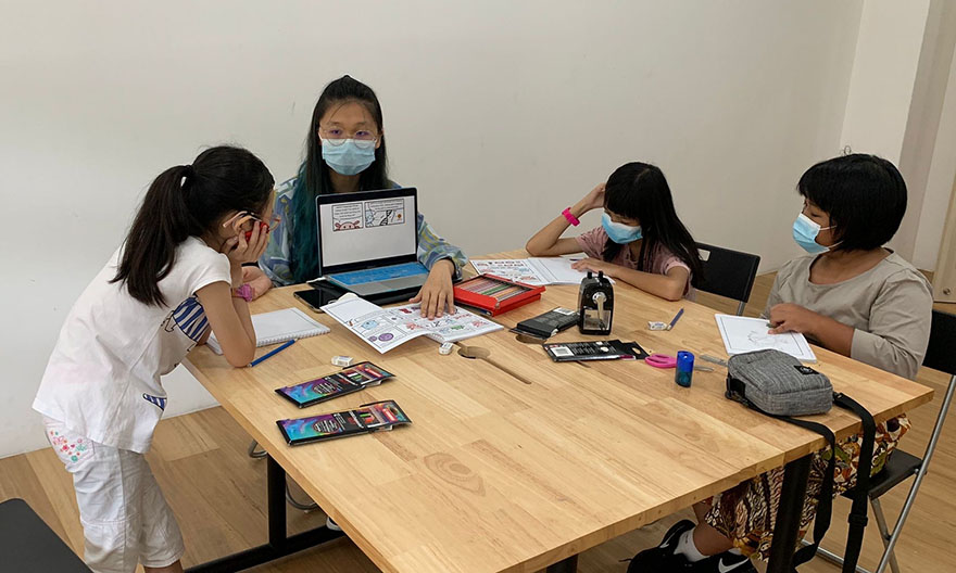 Huanyan, from the Class of 2023, is sitting down on a chair in front of the table full of notebooks, drawings, and stationaries with three students around where she holds a laptop with comic artwork on display, facing the students at the table where the students listen.