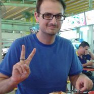 A headshot of smiling Andrew M Bailey who has short brown hair and is wearing a blue t-shirt and black-rimmed glasses. He is sitting with his food at a hawker centre, posing with the peace hand sign.