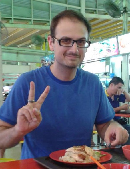 A headshot of smiling Andrew M Bailey who has short brown hair and is wearing a blue t-shirt and black-rimmed glasses. He is sitting with his food at a hawker centre, posing with the peace hand sign.