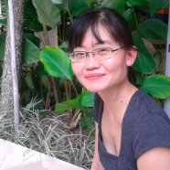 A headshot of smiling Claudine Ang who has short black hair with bangs, wearing a grey top and glasses with green plants in the background.