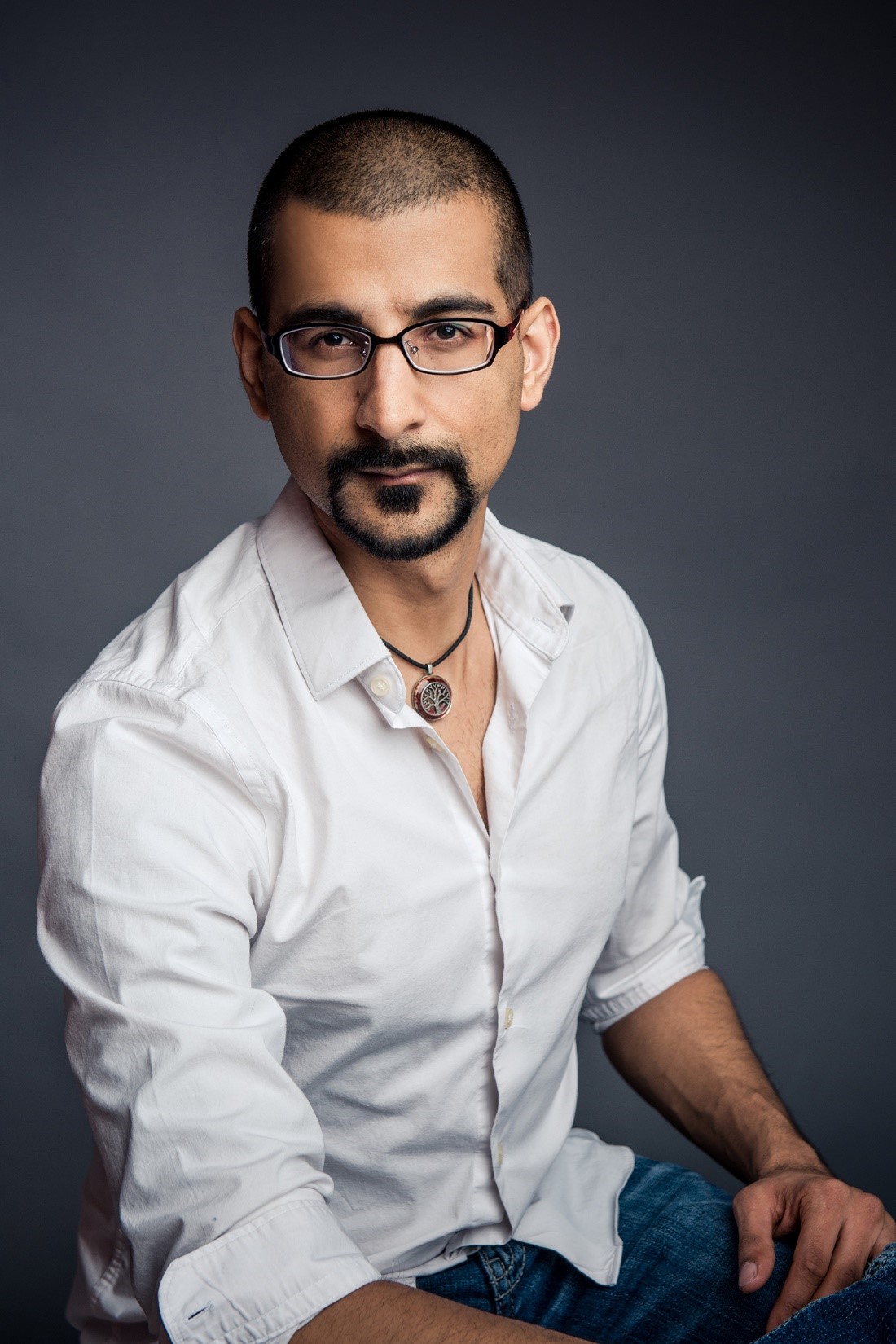 A headshot of smiling Neil Mehta who has buzz-cut black hair, a mustache, and a black beard. He is wearing a white collared shirt, glasses, and a black necklace with a pendant.