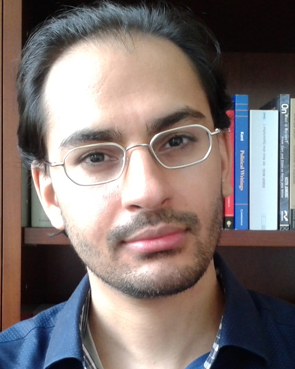 A headshot of smiling Taran Kang who has short black hair, a mustache, and a beard. He is wearing glasses and a dark blue collared shirt. Behind him is a bookshelf filled with books.