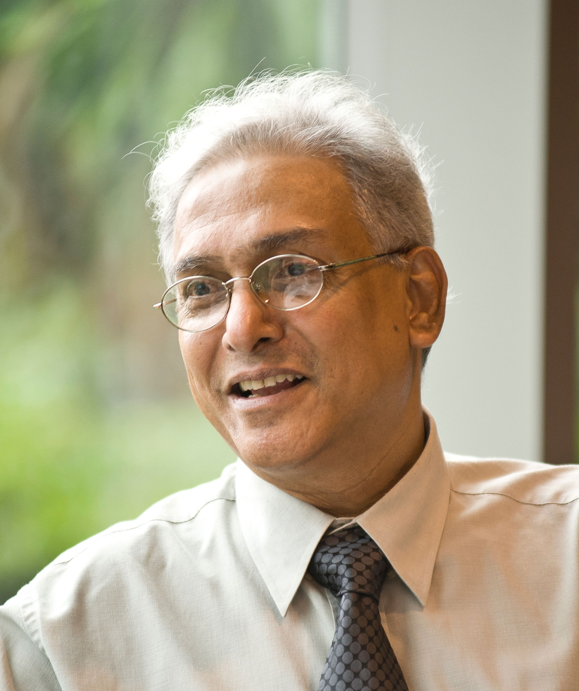A headshot of smiling Rajeev S Patke who has short grey hair, wearing glasses, a white collared shirt, and a black tie. He is sitting in front of a large window that overlooks greenery in the background.