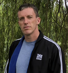 A headshot of smiling Steven Green who has short brown hair. He is wearing a black Yale-NUS jacket with a blue t-shirt underneath the jacket. He is standing in front of green trees.