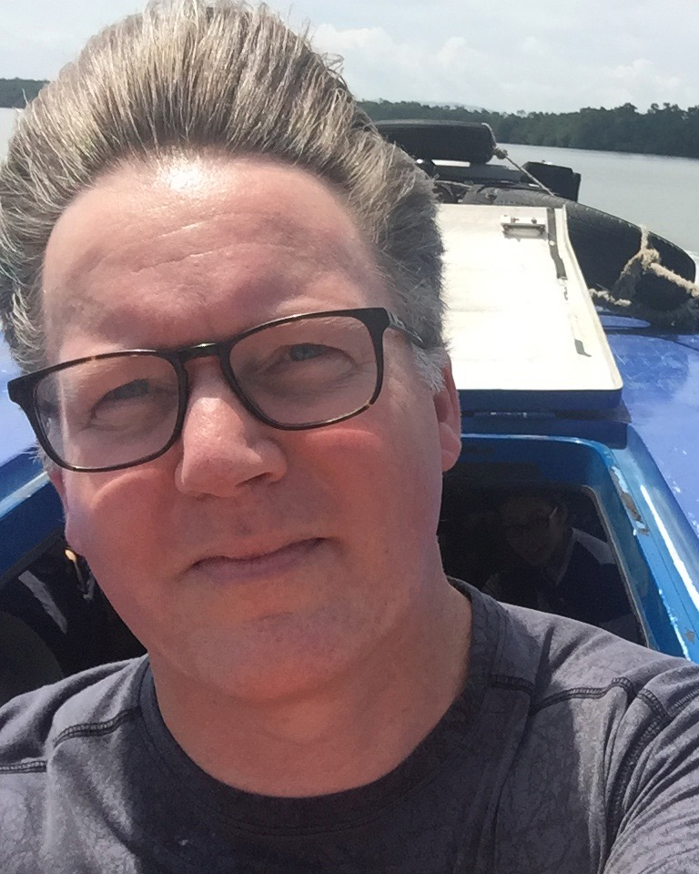A headshot of smiling Philip Johns in a boat, his light brown hair pushed back by the wind. He is wearing glasses with a black frame and a grey T-shirt.
