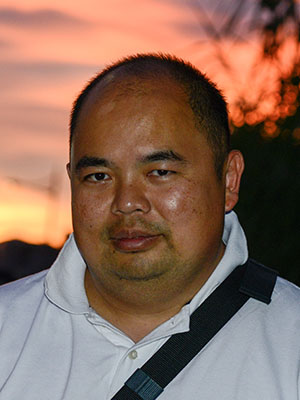 A headshot of smiling Eugene Choo who has short black hair, wearing a white polo shirt, and a cross-body bag. He is standing in front of a vibrant sunset with streaks of yellow, orange, and violet in the sky.