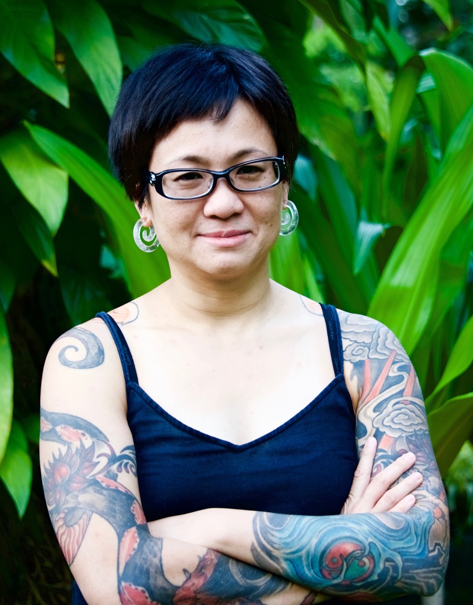 A headshot of smiling Lynette J Chua who has short black hair with pixie haircut, wearing glasses with a black frame and translucent earrings in a spiral shape. She is wearing a black tank top, showing a full tattoo sleeve on her left arm depicting roiling oceans and clouds. A large tattoo of an intricate red and black dragon coils around her right arm.
