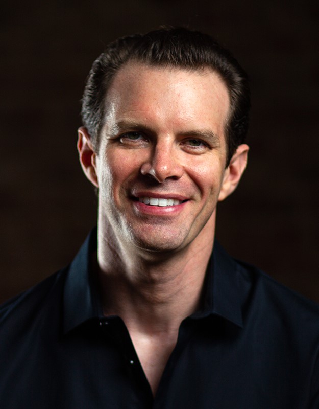 A headshot of smiling Jonathan Vandenberg who has brown hair which is combed back. He is wearing a black collared shirt.