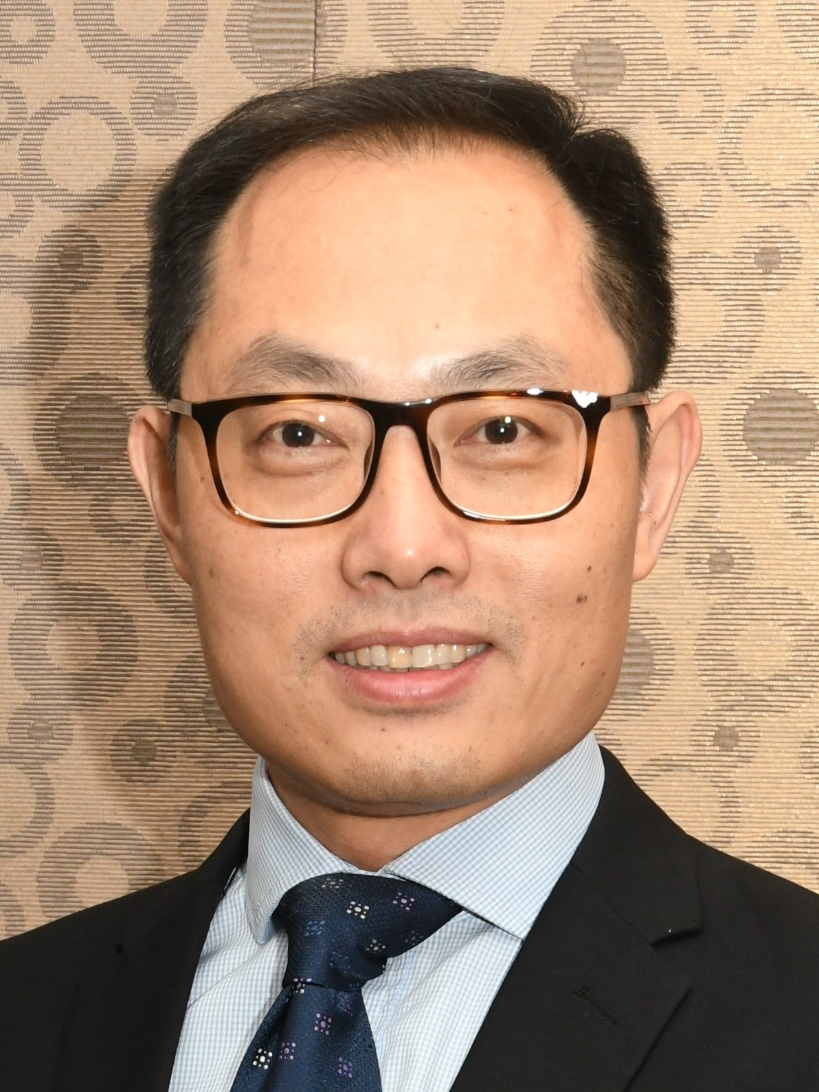 A headshot of smiling Zhang Jianlin who has short black hair. He is wearing glasses with a black frame, a black suit with a light blue collared shirt, and a navy-blue tie.