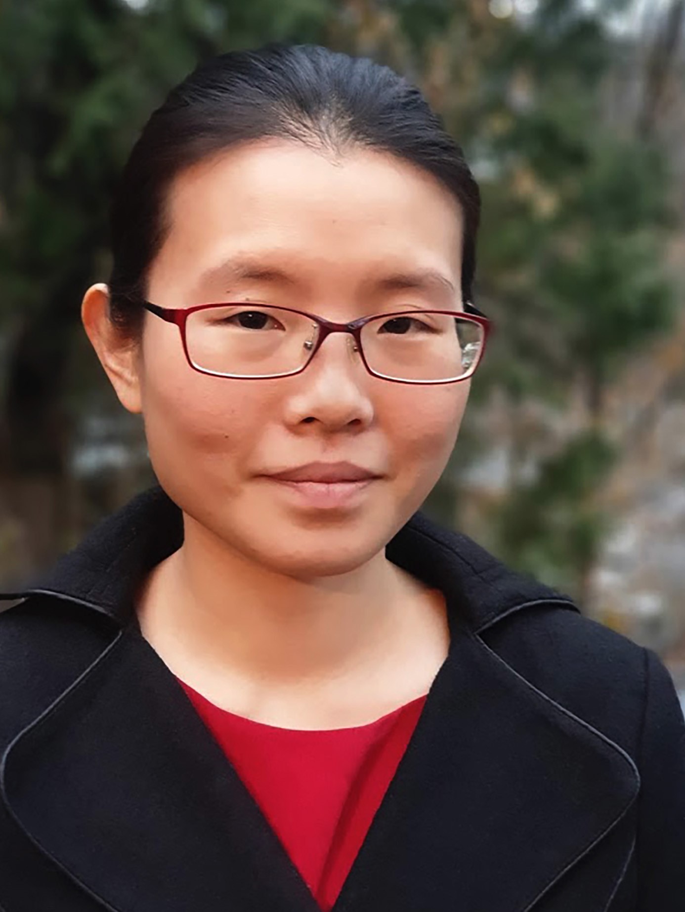 A headshot of Lau Ting Hui who has black hair which is tied back. She is wearing glasses with a red frame and a black jacket with a red top underneath. She is standing in front of greenery.