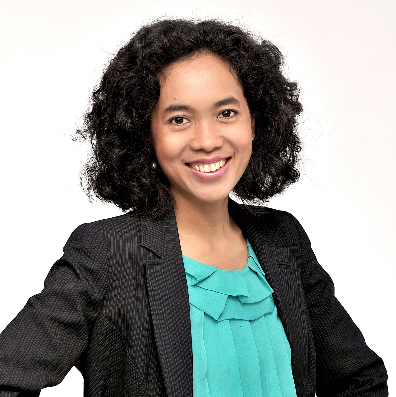 A headshot of smiling Naila Maya Shofia who has curly black hair at shoulder length. She is wearing a black blazer with a bright turquoise blouse.