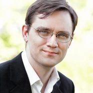 A headshot of smiling Michael D. Adams who has short brown hair, wearing a black suit and glasses with greenery in the background.