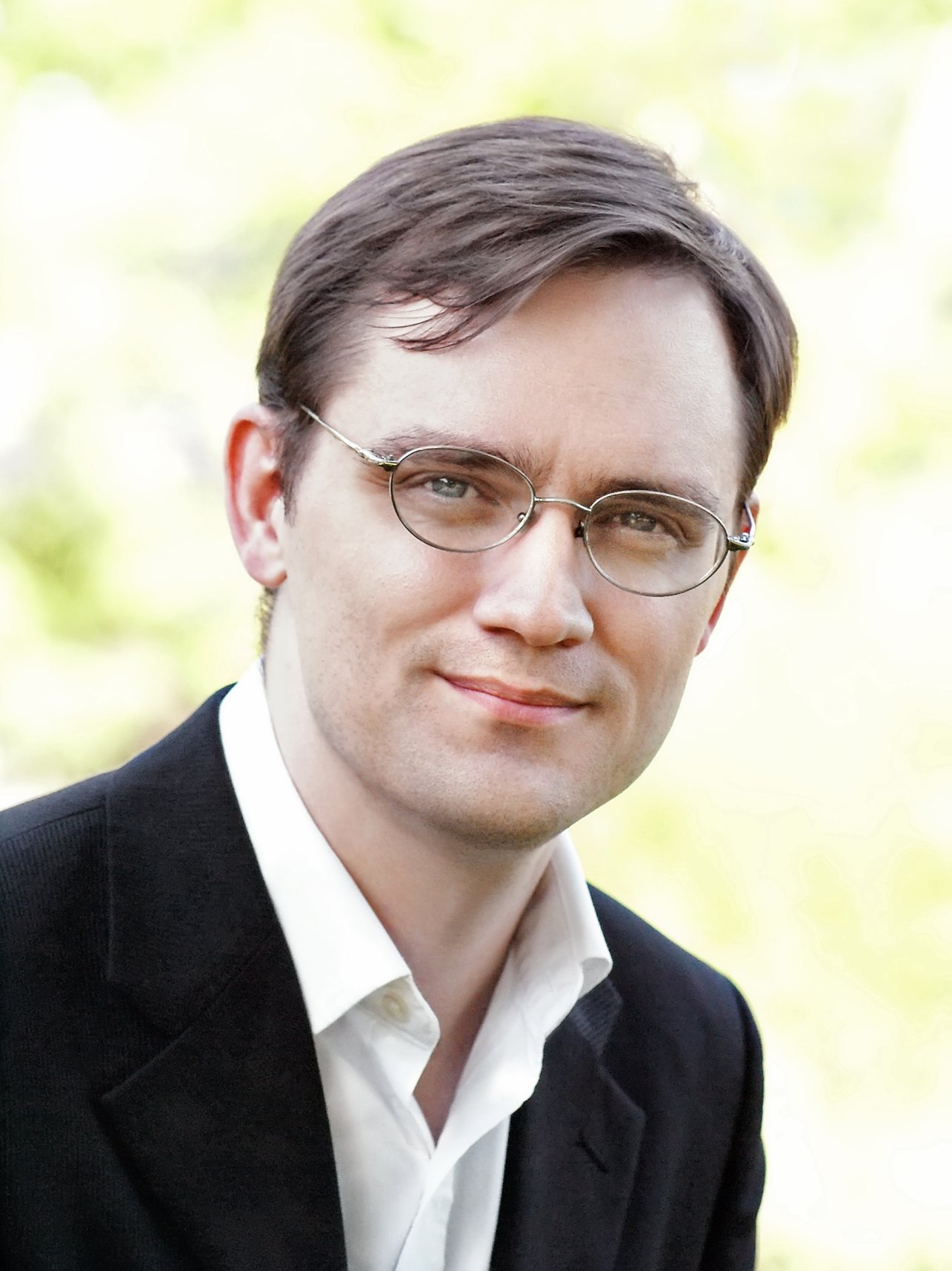 A headshot of smiling Michael D. Adams who has short brown hair, wearing a black suit and glasses with greenery in the background.
